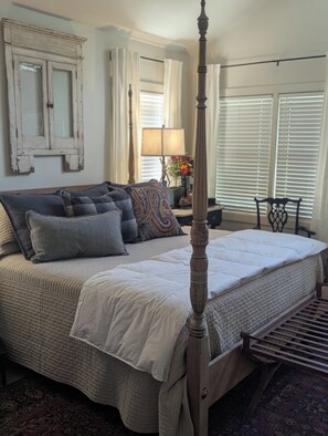 A bedroom with a four-poster bed, a nightstand with a lamp, a chair, and a window with blinds.
