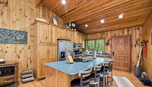 Interior de una cabaña de madera con área de cocina, taburetes de bar y una puerta grande de madera.