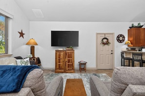 A living room with a flat-screen TV, wooden cabinets, a small table, and a door with a wreath. - 4-BD/2-BA Southern Retreat with Waterfront Patio (Gainesville)