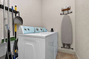 A laundry room with a front-loading washing machine, ironing board, and various cleaning tools. - 4-BD/2-BA Southern Retreat with Waterfront Patio (Gainesville)