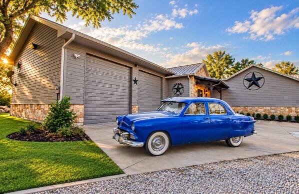 Een blauwe oldtimer geparkeerd voor een huis met een garage, een sterlogo op de garage deur, en een klok aan de muur.
