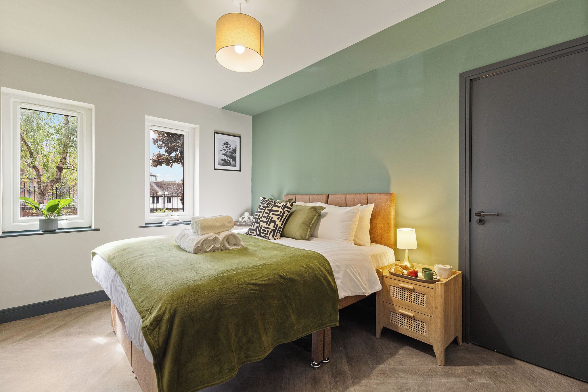 Une chambre à coucher avec un lit, une table de chevet, une lampe et une fenêtre donnant sur des arbres.