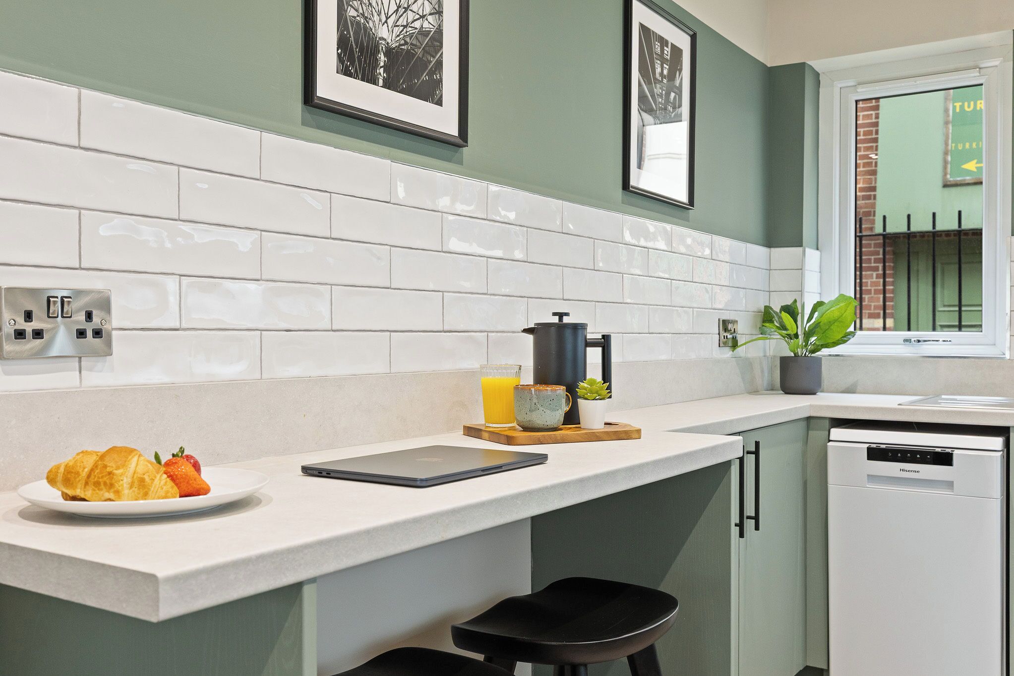 Uma cozinha moderna com azulejos brancos, cafeteira, tábua de frutas, laptop e lava-louças.