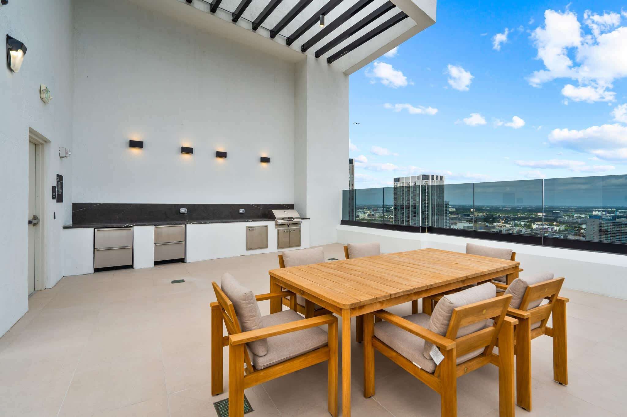 A rooftop terrace with a wooden table and chairs, a built-in grill, and a city view.