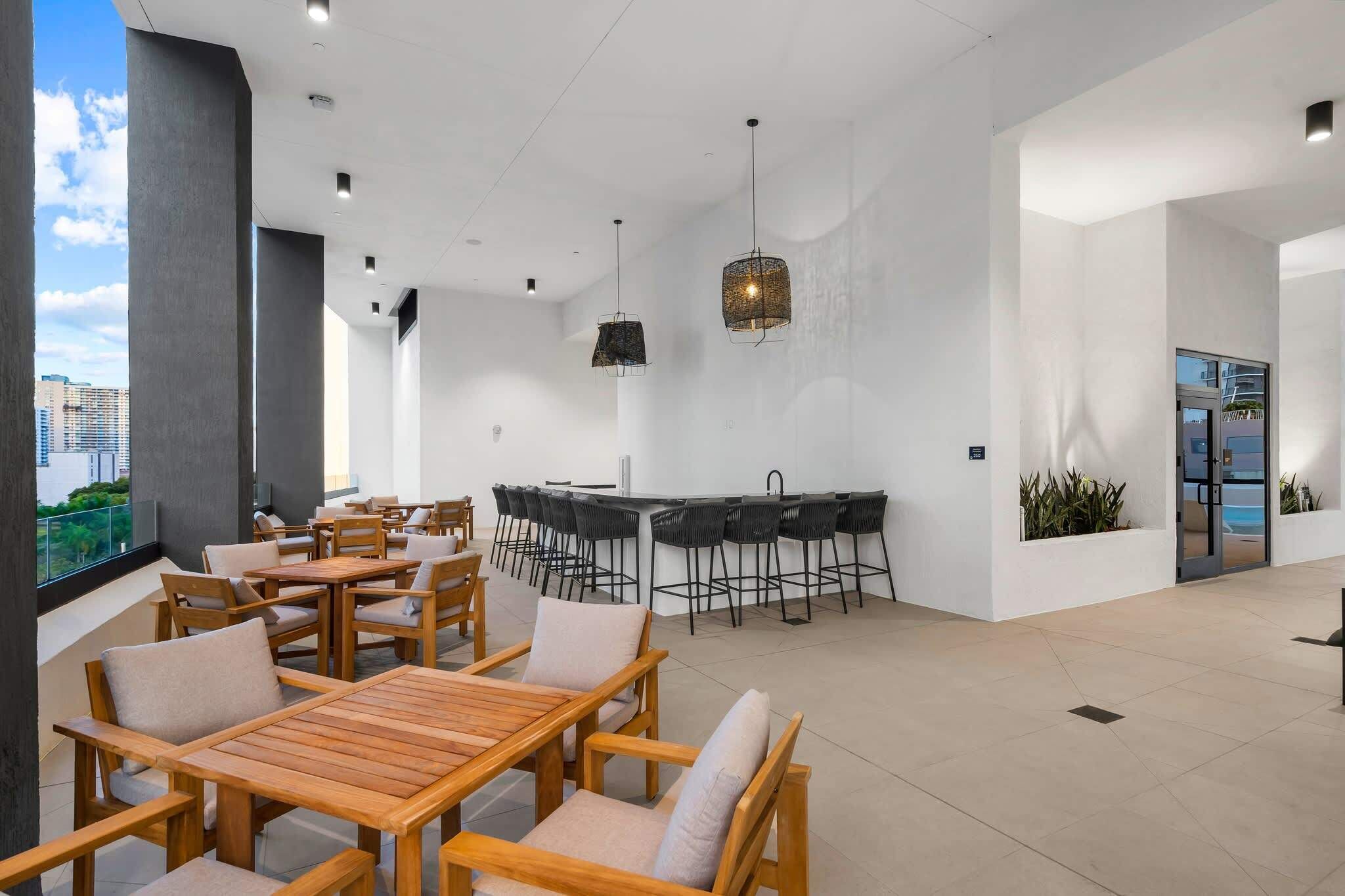 A modern indoor dining area with wooden tables and chairs, a bar area with stools, and a view of the city through large windows.