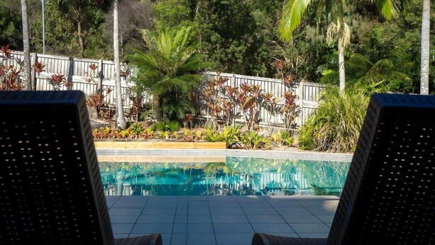 A swimming pool with tiled flooring, surrounded by a wooden fence and various plants.