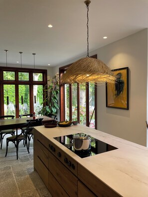 A modern kitchen with a central island, wooden cabinets, and a large window providing a view of greenery. - Unique London family-sized home with stunning Natural Pool (London)