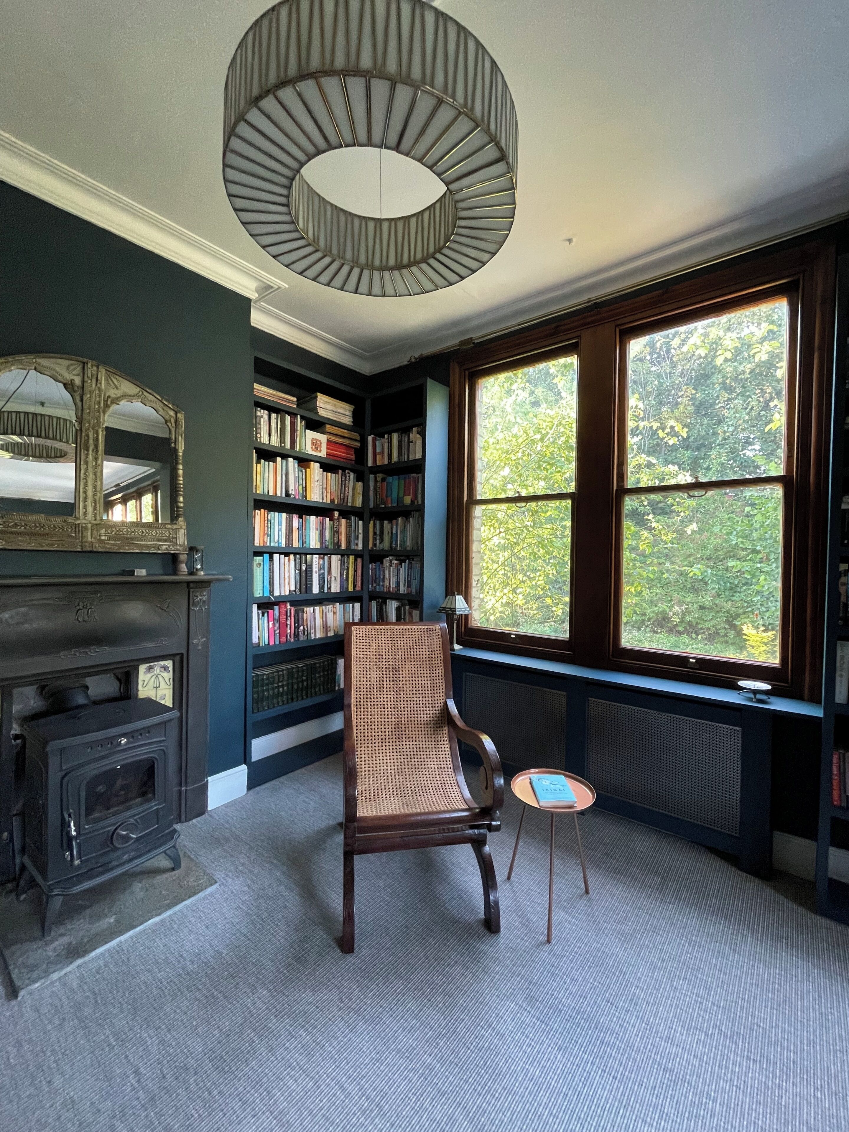 A room with a fireplace, a bookshelf, a chair, a small table, and a large window with a view of trees.