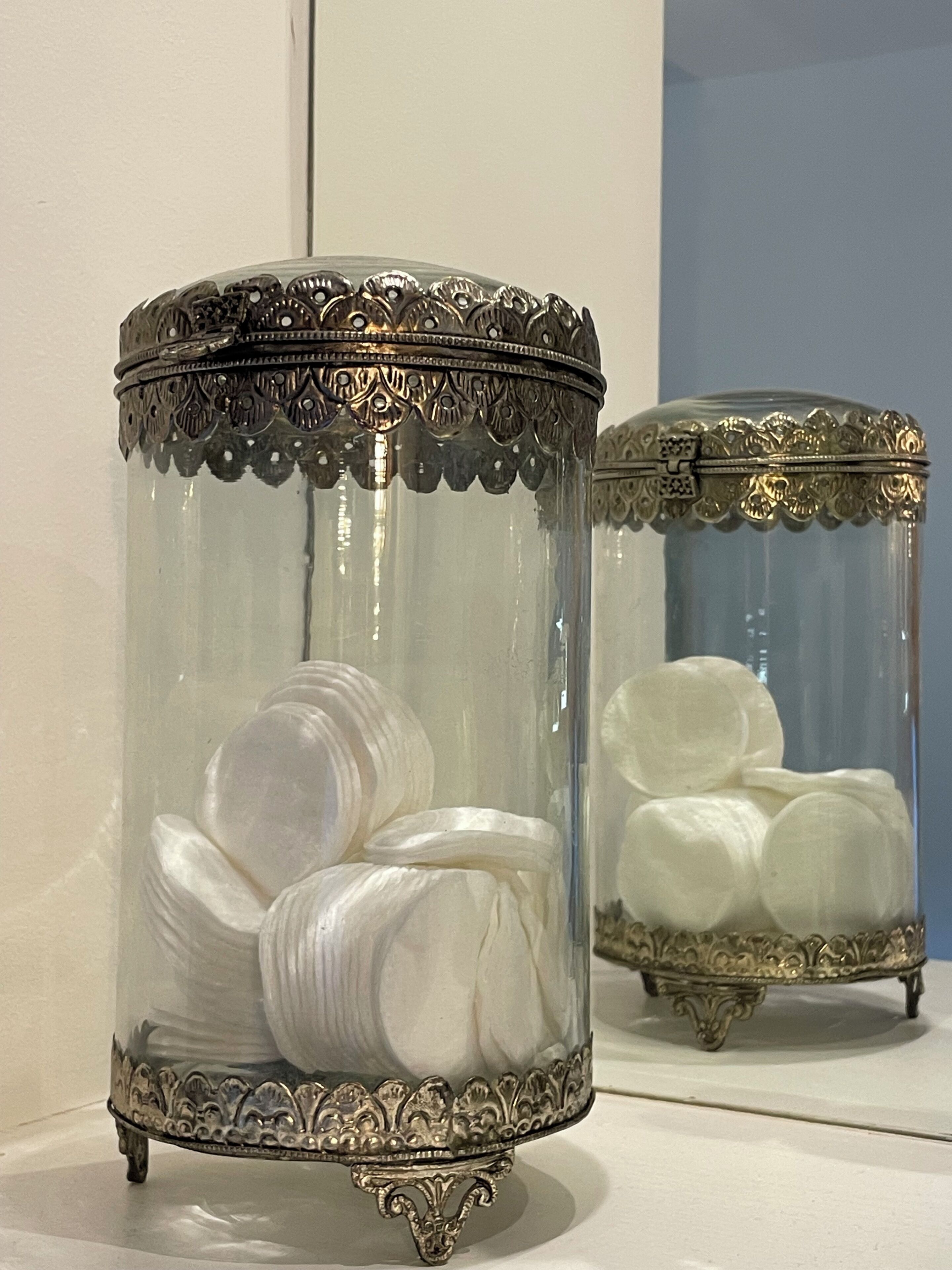 Two ornate glass containers with metal bases, each holding rolled white fabric.