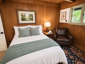 A bedroom with a bed, a nightstand, a lamp, a leather chair, and a window with a view of trees. - Boutique Riverside Retreat (Seaford)