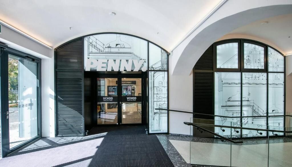A modern building entrance with a glass facade, a large 'PENNY' sign, and architectural drawings on the glass.
