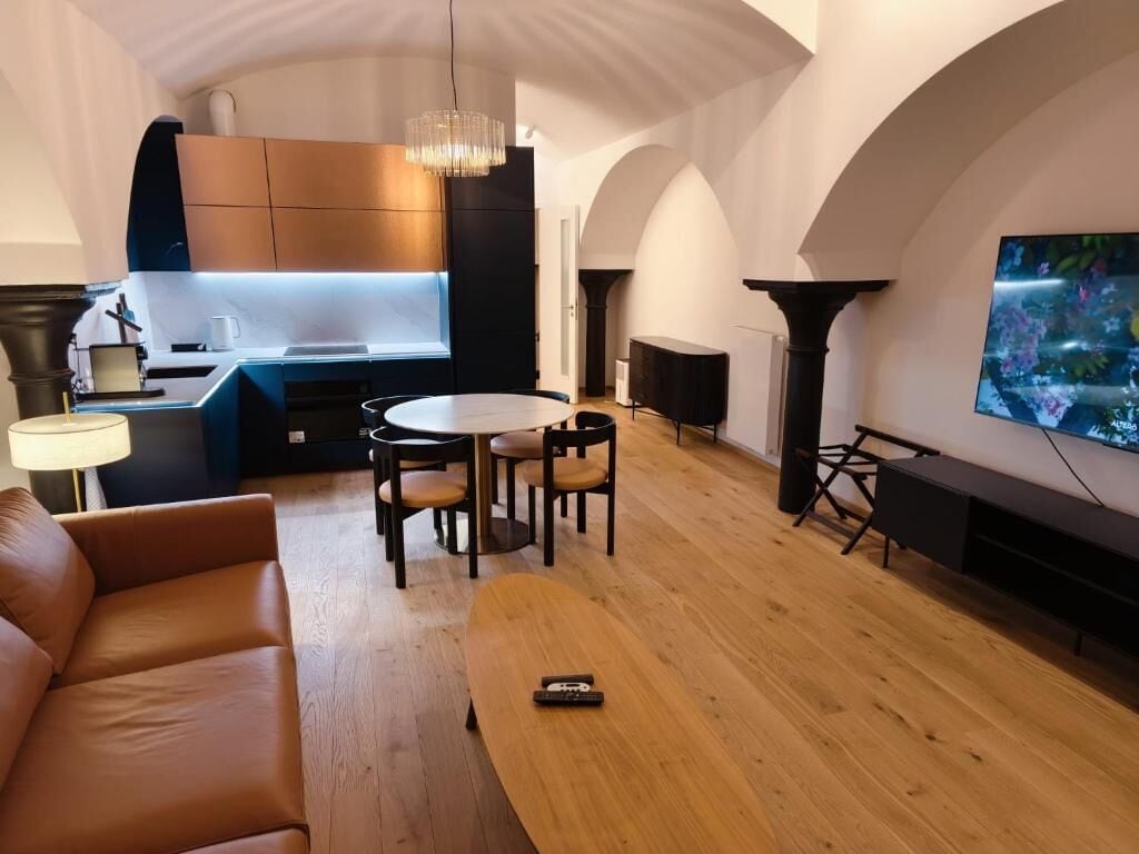 A modern living room with a sofa, a dining table with chairs, a TV, and a kitchen area with cabinets and a pendant light.