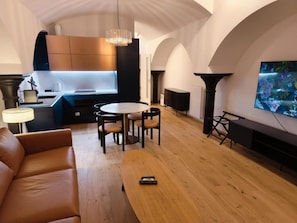 A modern living room with a sofa, a dining table with chairs, a TV, and a kitchen area with cabinets and a pendant light. - Free Parking, Nuslesky Pivovar Blue Arch Luxury Apartment With Garden (Prague)