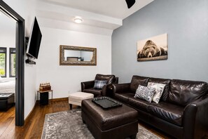 A modern living room with a leather sofa, a coffee table, a flat-screen TV, a mirror, and a painting on the wall. - Frisco Retreat | Creekside • Unbeatable Location (Frisco)