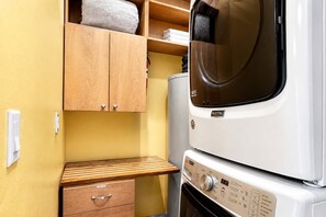 A compact laundry room with a stacked washer and dryer, wooden cabinets, and a small wooden bench. - Frisco Retreat | Creekside • Unbeatable Location (Frisco)