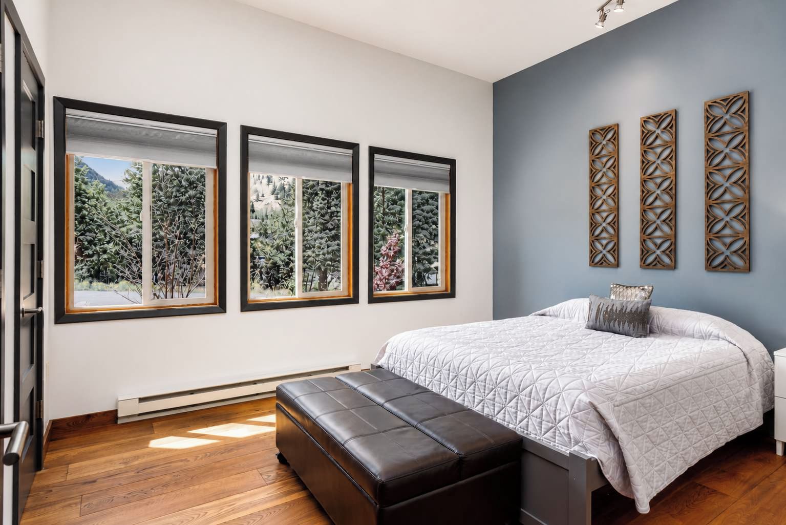 A bedroom with a bed, a bench, and three windows with a view of greenery.