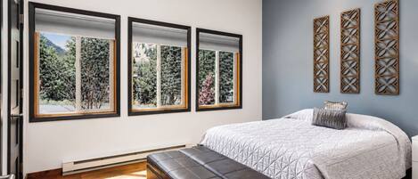 A bedroom with a bed, a bench, and three windows with a view of greenery.