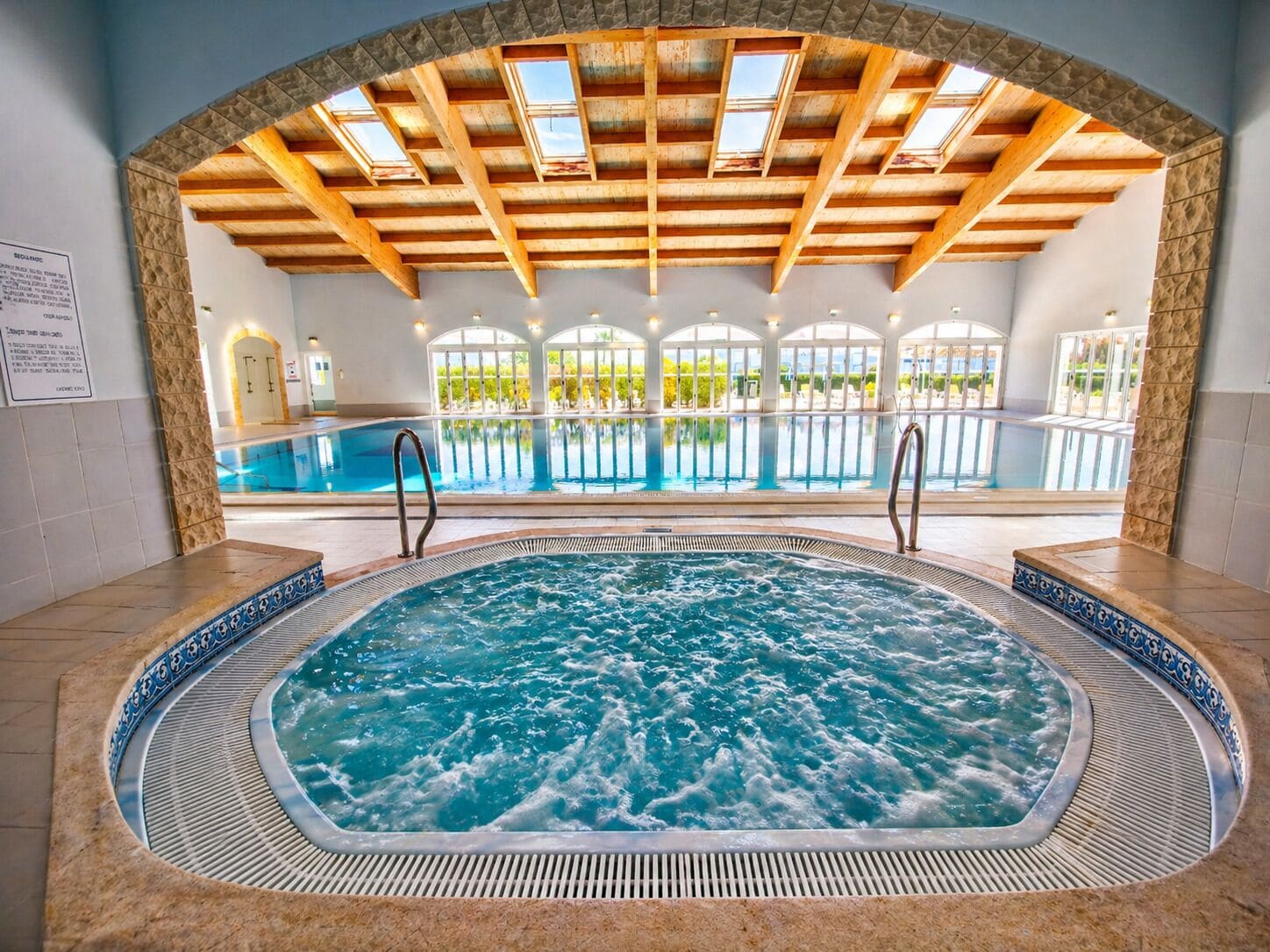 En indendørs swimmingpool med jacuzzi, træbjælker og udsigt til en udendørs pool.