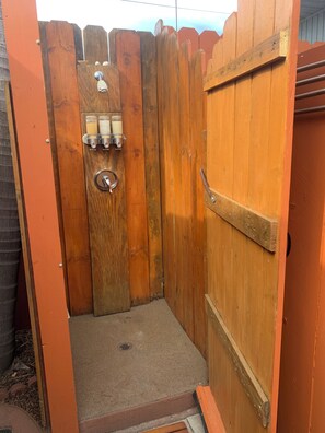 Une douche extérieure en bois avec une porte en métal, une petite étagère avec des bougies et un drain en métal.
