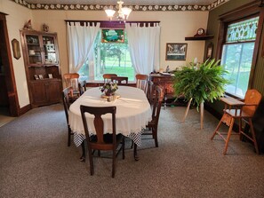 Uma sala de jantar com uma mesa redonda, cadeiras, um candelabro, um armário e uma grande janela com vista para o exterior.