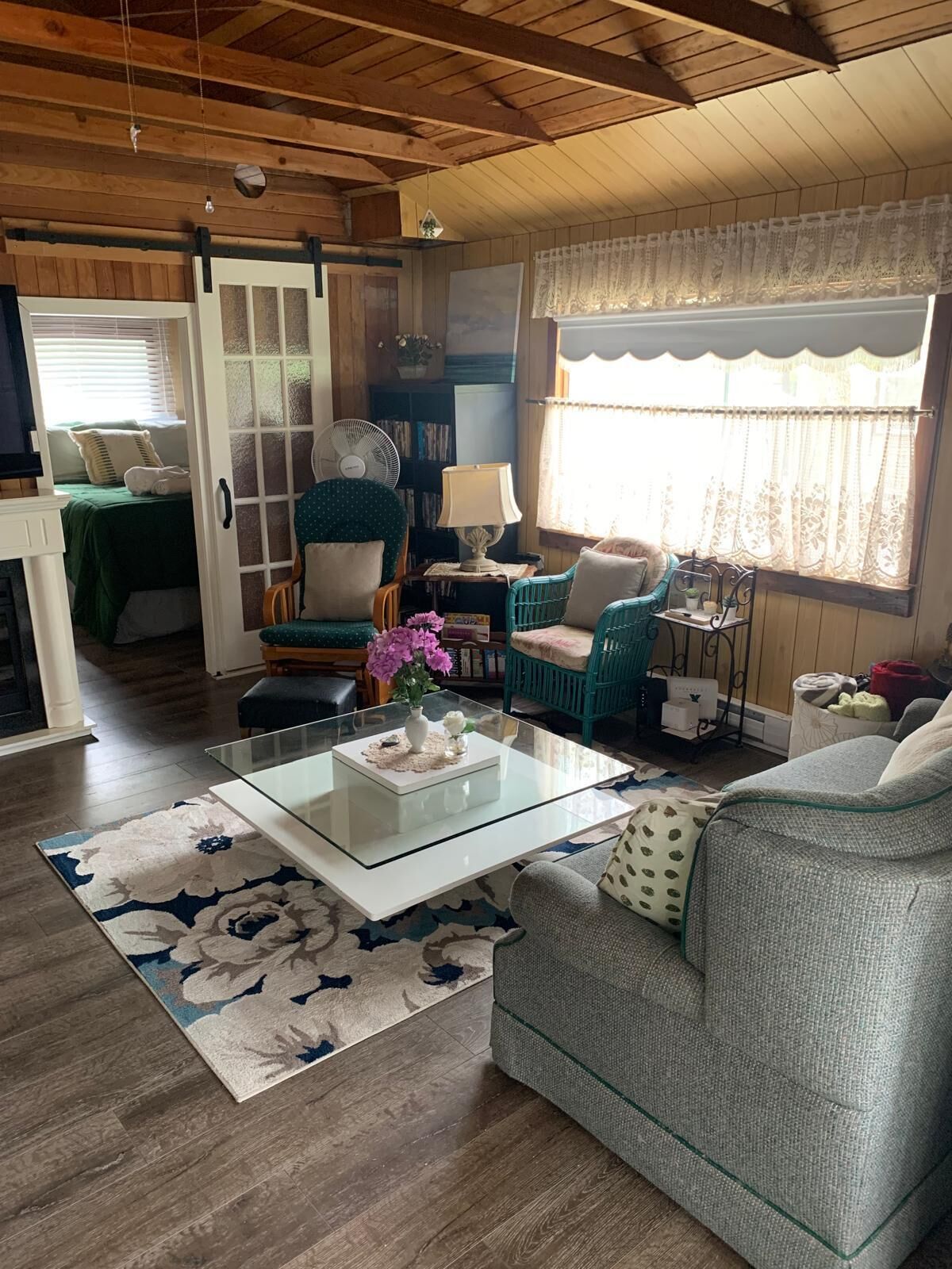 A cozy living room with a glass coffee table, a sofa, and a chair. There is a bookshelf and a window with curtains.