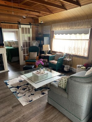 A cozy living room with a glass coffee table, a sofa, and a chair. There is a bookshelf and a window with curtains.