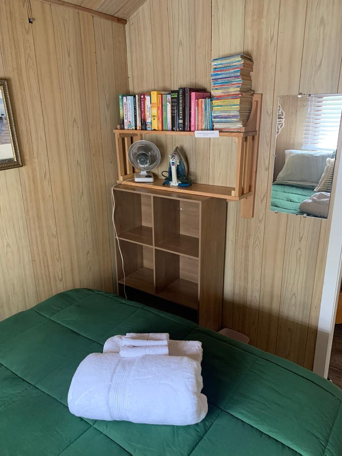 A room with a green bed, a wooden shelf with books, a fan, and a mirror reflecting a bed.
