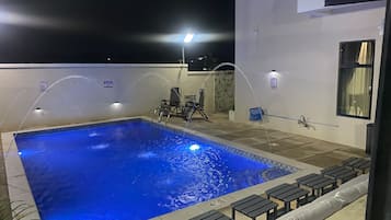 Una piscina rectangular con borde de azulejos, rodeada de sillas de descanso y una pequeña mesa, bajo una zona techada.