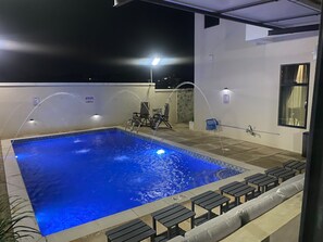 A rectangular pool with a tiled edge, surrounded by lounge chairs and a small table, under a covered area with a roof. - Deval Den (Enugu)