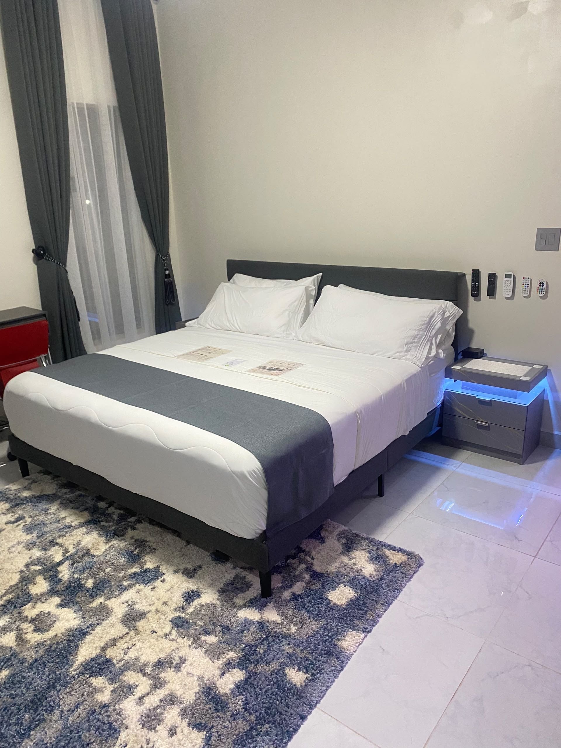 Una camera d'albergo moderna con un letto grande, un comodino con una lampada, un tavolino e un pannello di controllo a parete.