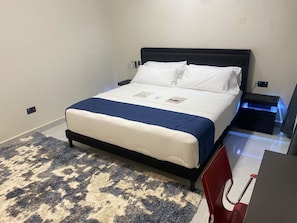 A hotel room with a bed, a nightstand, a desk, and a chair. - Deval Den (Enugu)