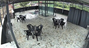 A covered outdoor seating area with tables and chairs, a glass partition, and a view of greenery. - Deval Den (Enugu)