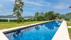 Une piscine à l’eau d’un bleu cristallin, entourée d’une pelouse bien entretenue et de palmiers, avec une vue sur un lac et des collines en arrière-plan.