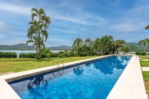 A swimming pool with clear blue water, surrounded by a well-maintained lawn and palm trees, with a view of a lake and hills in the background. - Pacifico Azul Punta Plata 517 (Potrero)