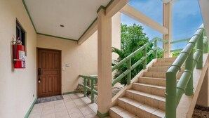 Un hall d’hôtel avec une porte en bois, un extincteur, un escalier avec des rampes vertes et une vue sur la verdure à l’extérieur.