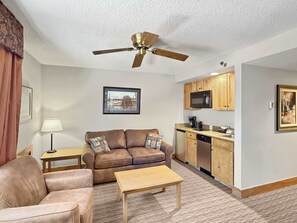 A compact living space with a sofa, a coffee table, a ceiling fan, a kitchen area with wooden cabinets, and a framed picture on the wall. - Grand Lodge 259: Ski In/Ski Out 2BR, 2BA, Sleeps 6 - Pool, Hot Tub, Dogs OK (Crested Butte)