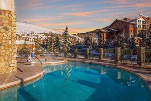A pool area with a stone wall, a hot tub, and a view of a residential area with snow-covered roofs and trees. - Rare Grand Lodge 2 Bedroom, perfect for Families — Pet Friendly, Walk to the Lifts, Pool & Hot Tub (Crested Butte)