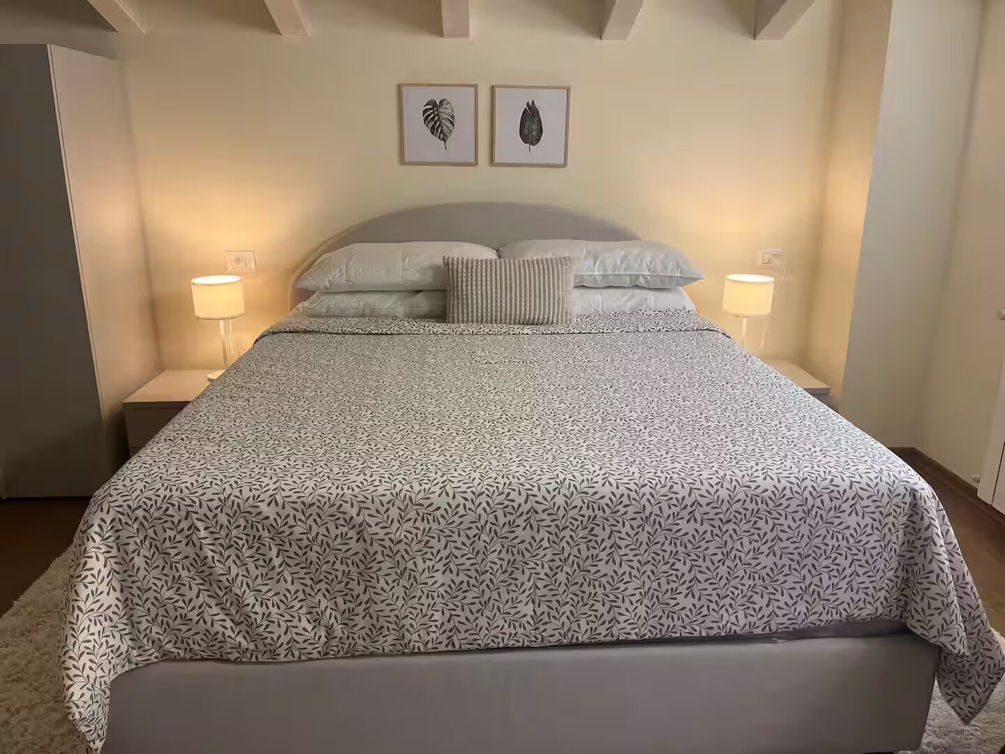 Un letto rifatto con cura, una coperta a motivi decorativi, due opere d'arte con foglie incorniciate a parete e lampade ai lati del letto.