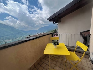 A balcony with a yellow table and chair, a potted plant, and a view of mountains and a lake.