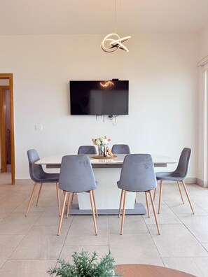 A modern dining area with a flat-screen TV mounted on the wall, a round table with chairs, and a decorative vase with flowers.