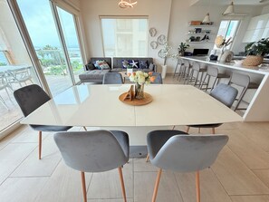 A modern dining area with a large table, grey chairs, and a view of the ocean through large windows.