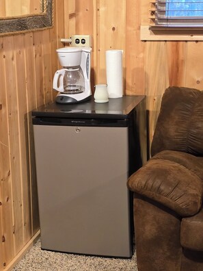 Ein kompakter Kühlschrank mit Kaffeemaschine und Papierhandtuchhalter auf der Oberseite, ein braun gepolsterter Stuhl und Holzpaneele.