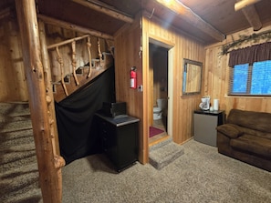 A wooden interior with a staircase, a desk, a small kitchenette, a couch, and a window with blinds.