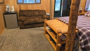 Ein gemütliches Blockhäuschen im Inneren mit einer Couch, einem kleinen Kühlschrank, einer Holzbank und einem Bett mit karierter Bettwäsche.