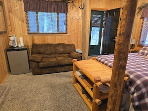 Ein gemütliches Blockhäuschen im Inneren mit einer Couch, einem kleinen Kühlschrank, einer Holzbank und einem Bett mit karierter Bettwäsche.