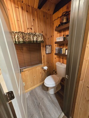 Une salle de bain en bois comprenant des toilettes, une fenêtre avec des stores, une étagère avec des articles de toilette et un panneau décoratif encadré.