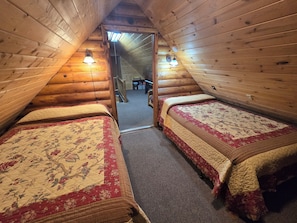 Une cabane avec deux lits, des murs en bois, un plafond en pente et un escalier apparent menant à un niveau supérieur.