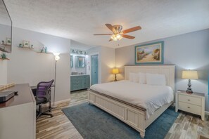 A bedroom with a bed, a desk, a chair, a ceiling fan, a painting, and a bathroom visible through an open door. - Turtle House - Quiet 2BR 2BA - Seconds from beach, short walk to Pier Park. (Panama City Beach)