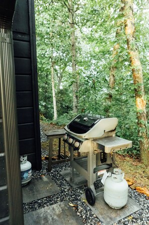 An outdoor grill setup with a gas cylinder and a portable table, surrounded by a forested area. - Azalea Ridge Retreat - Cozy family cabin near Dahlonega (Dahlonega)