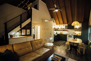 En rummelig opholdsstue med en lædersofa, et spisebord med stole, et køkken med moderne hårde hvidevarer, og en trappe med gelænder.